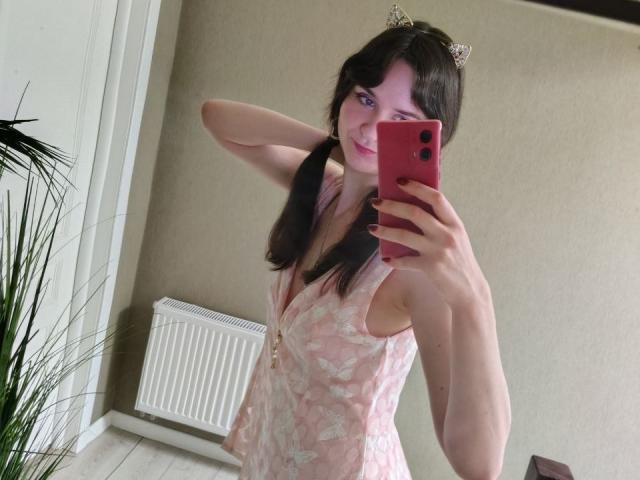 BerryDiva with long brown pigtails, wearing a pink butterfly dress, cat ears, holding a red phone, smiling, showing small tits, standing in a mirror selfie.