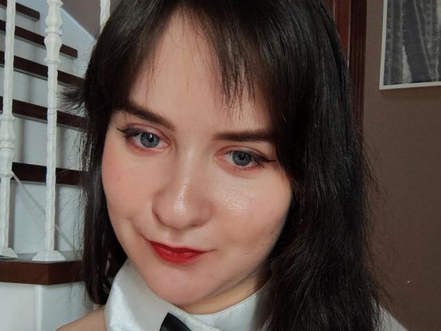 BerryDiva has fair skin, blue eyes, black hair, and red lipstick. She's wearing a white shirt with a black tie, looking cute with a slight smile.