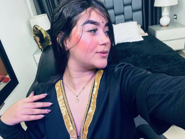 AnaShalimarr mit dunklem Haar, rosiger Wangen und einem schwarzen Hemd mit goldener Verzierung, das ihre Brüste betont, wobei sie eine Hand auf ihrer Brust hat, in einem Schlafzimmer. Sie trägt eine Halskette..