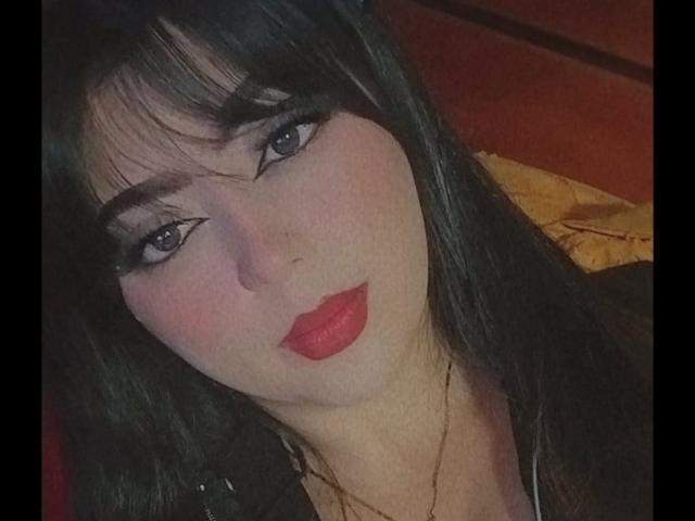 NathyHeat has dark black hair, red lipstick, and black eyeliner with long lashes, looking directly at the camera with a slight pout, wearing a black top.