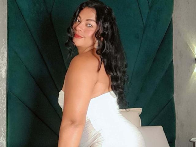 BrunetteScarlet with curly black hair, smiling, looking over her shoulder, wearing a strapless white dress, showing her bare back and white butt, standing in a dark green room.