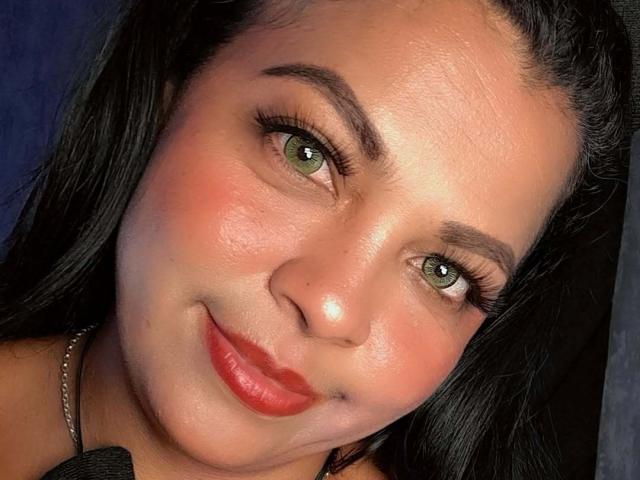 BrunetteScarlet has green eyes, red lipstick, and a slight smile, with black hair and a shiny face. Her nose has a small, visible blemish. She's wearing a silver necklace.
