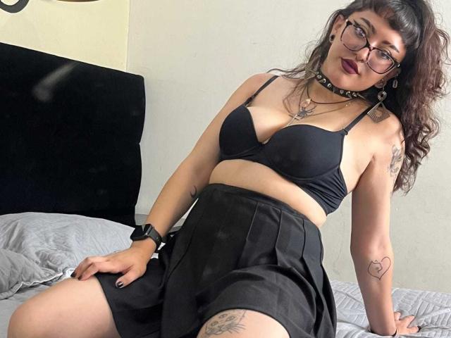 EmilyAmatista with dark curly hair, glasses, black choker, black bra, black skirt, tattoos, sitting on bed, hand on thigh, black wristwatch, looking at camera, red lipstick.