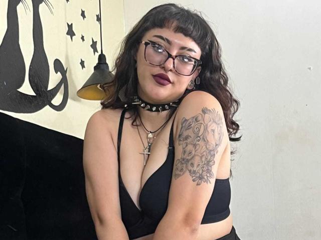 EmilyAmatista with dark wavy hair, glasses, and deep red lipstick, wearing a black bra and spiked choker, showing a tattoo on her right arm, and a cross necklace.