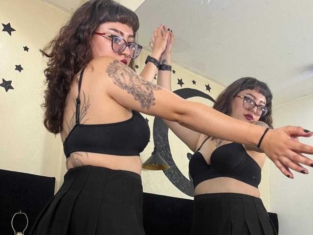 EmilyAmatista with curly brown hair, glasses, black bra, and skirt, tattoo on arm, mirror reflection, black nail polish, and wrist bracelet, stretching in front of a mirror.
