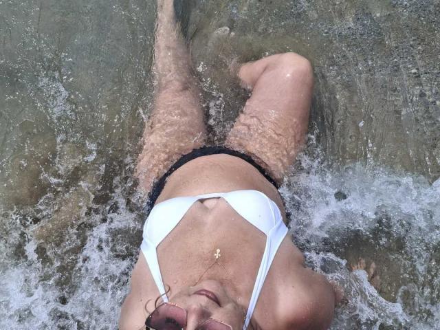 BozhenaNude lying in shallow water, white bikini top, black bikini bottoms, arms above head, wet skin, small gold necklace, legs slightly apart, water splashing around her.