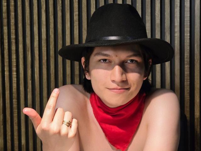 ErossGuy with pale skin, black hat, red scarf, and ring on index finger, giving a "rock on" hand gesture, smiling slightly.