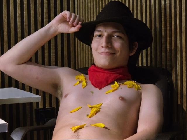 ErossGuy, shirtless, wearing a black hat and red bandana, with yellow banana petals on his chest, sitting, smiling, right arm raised, holding hat, in a chair.
