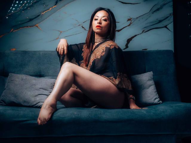 AngelinaBlanche with long brown hair, red lipstick, black lace robe, and exposed pussy, sitting on blue sofa, legs crossed, confident expression, dark background, soft lighting.