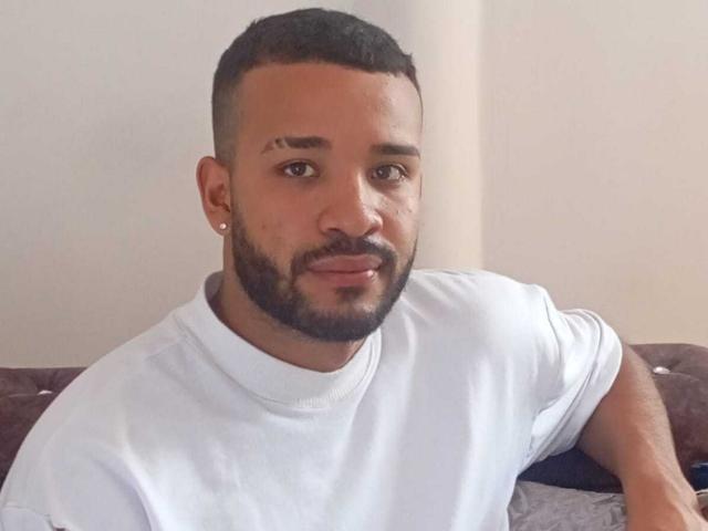 BrayanHotLover, a dark-skinned dude with a short beard and undercut, wearing a white turtleneck, has a small scar above his right eyebrow and a diamond earring.