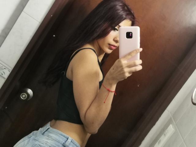 DanikaSerov with long dark hair, wearing a black crop top and light blue jeans, taking a mirror selfie with a pink phone, showing her butt and red bracelet.