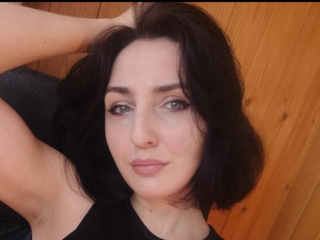 GertieDi has dark, wavy hair, green eyes, and a slight smile. She's wearing a black top, with one arm raised, showing her armpit. Wood panel background.