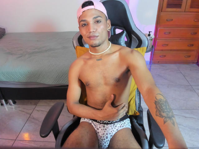 AlejoMorgan, shirtless, wearing a pink backward cap, white pearl necklace, and white polka-dot boxer briefs, giving a thumbs-up with a tattooed arm, sitting in a gaming chair.