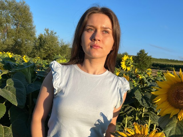 AlvaGreer with medium tits, wearing a white sleeveless top, standing in a sunflower field, blue sky, brown hair, green eyes, slight smile, sunlight, clear day.