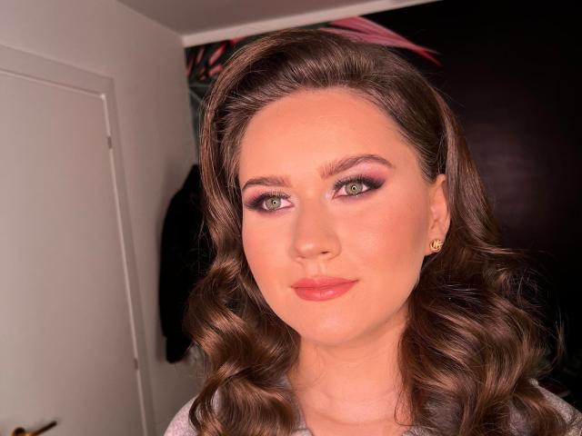 AlvaGreer has wavy brown hair, green eyes, and pink eyeshadow. She's wearing pink lipstick, and has a small gold earring on her right ear. She's looking at the camera.