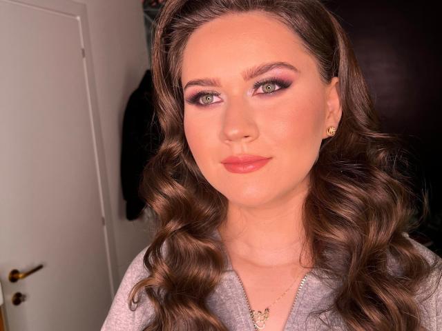 AlvaGreer has wavy brown hair, pink eyeshadow, and glossy pink lips. She's wearing a grey zip-up and small gold earrings. Her tits are partially visible.