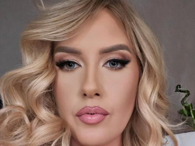 OndinaNoir has blonde, wavy hair, green eyes with thick black eyeliner, and pink glossy lips. Her skin is smooth and fair. She's looking directly at the camera.