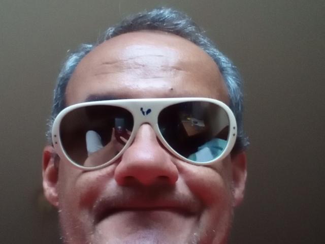 HotDrew's face with gray hair, wearing white sunglasses reflecting a mirror, smiling with a slightly wrinkled forehead and visible nose.