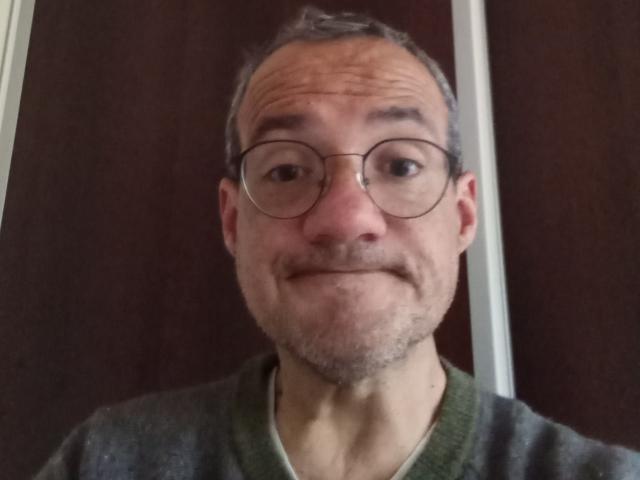 HotDrew has a goofy smile, thin mustache, and gray hair, wearing round glasses and a green V-neck sweater, with a slightly wrinkled forehead.