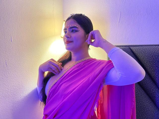 NayaYalitza wearing a pink sari with gold trim, white long-sleeve shirt, and red lipstick, has her hand near her face, looking to the side, with purple lighting.
