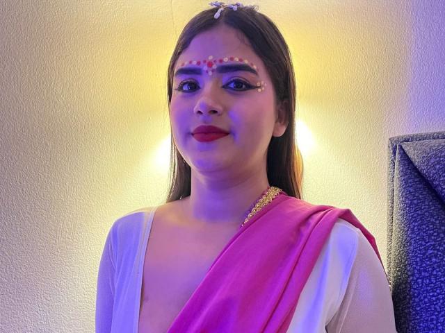 NayaYalitza has long brown hair, red lipstick, and white makeup dots on her forehead. She wears a pink and white sari with a gold necklace. She's smiling.