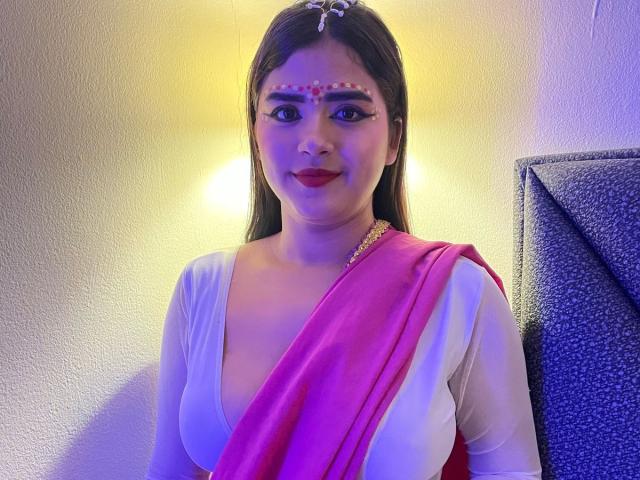 NayaYalitza with medium tits, wearing a pink and white sari, red makeup dots on her forehead, long brown hair, and red lipstick, smiling in a yellow-lit room.