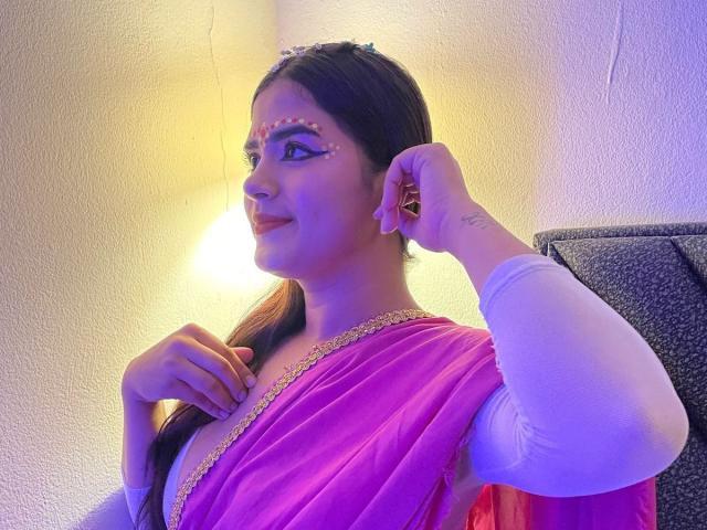 NayaYalitza with brown hair, wearing a pink sari, white long-sleeve top, and pink makeup on her face, smiling, raising her right hand, visible small tattoo on her wrist.