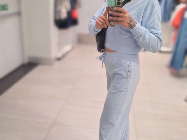 SammyDesire con una canottiera azzurra e pantaloni abbinati, mostra tatuaggi sulla mano, sta scattando un selfie allo specchio con il telefono, sfondo blu, pavimento piastrellato grigio, tiene i capelli neri..