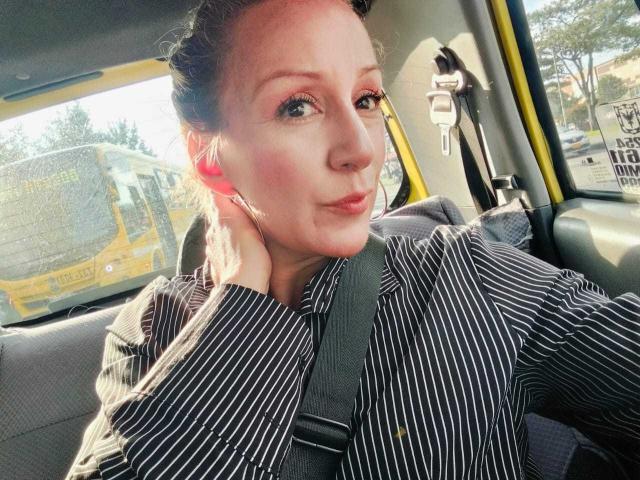 SammyDesire with puffy ass in striped shirt, seatbelt on, holding phone to ear, looking cute in car, yellow bus outside window. Sunlight on face, slight smile.