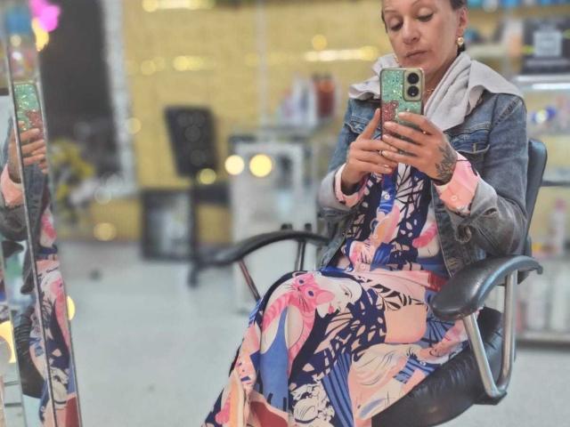 SammyDesire, con tatuajes, vistiendo una chaqueta vaquera y un vestido floral colorido, sentada en una silla, tomando un selfie en un espejo, sosteniendo un teléfono con una funda verde..