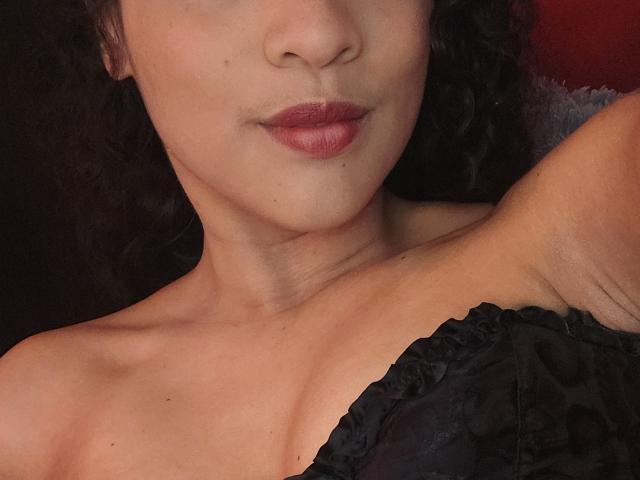 O rosto de Alejandra Montana, com batom vermelho escuro, cabelo cacheado preto e uma blusa de renda preta, que expõe a axila e os pequenos seios, mostrando um leve sorriso..