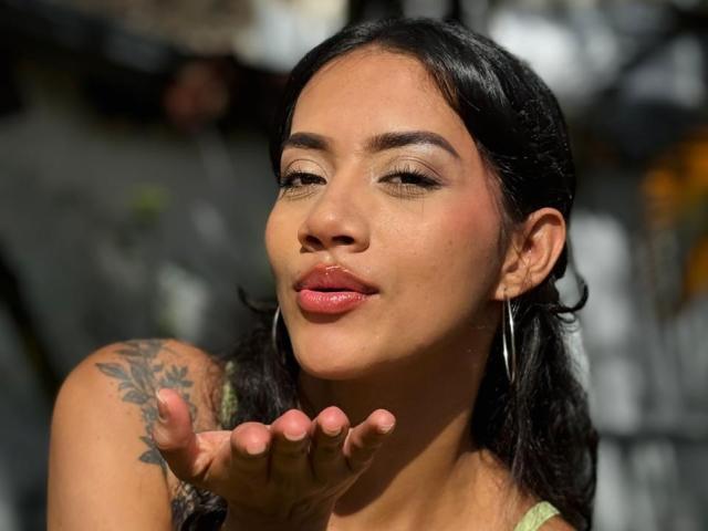 LunaGomezz mit dunklem Haar, Reifenohrringen und pinkem Lippenstift, die ihre Hand in die Nähe ihres Gesichts hält und dabei ein Tattoo auf ihrem linken Arm zeigt, lächelt mit geschlossenen Augen..