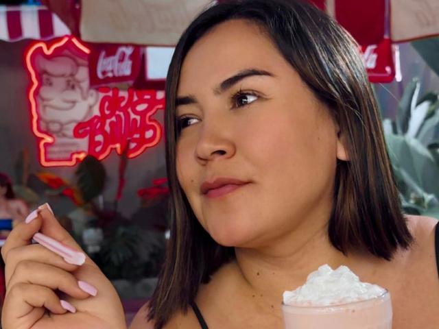 IsisClarkson with medium brown hair, holding a pink drink with ice, pink nails, and slight smile, looking to the side. Neon signs and plants in the background.