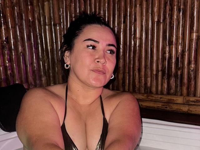 IsisClarkson with dark hair, wearing a brown halter top, showing her large tits, slightly puffy face, white ear gauges, and a confident, smirking expression, in a dimly lit room.