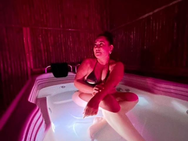 IsisClarkson sits in a hot tub, legs crossed, arms folded, wearing a black bikini, with red lighting highlighting her medium tits and dark hair in a bun.