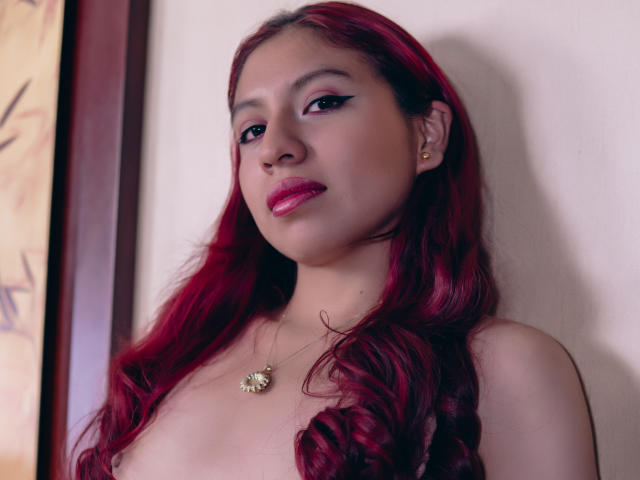 ZoeRedds has red, wavy hair, wearing a gold necklace with a sunflower pendant, and is topless, showing small, perky tits. She has pink lipstick and black eyeliner.