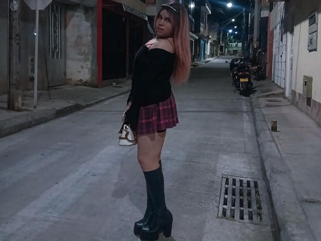EvelyneRosse with pink hair, black off-shoulder top, pink plaid skirt, black knee socks, and black platform boots, holding a white purse, standing on a dark street.