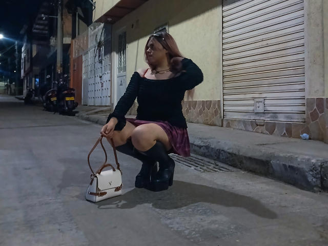 EvelyneRosse, squatting on a dark street, wearing a black off-shoulder top, pink skirt, black knee socks, and black boots, holding a white handbag, with her hand in her hair.