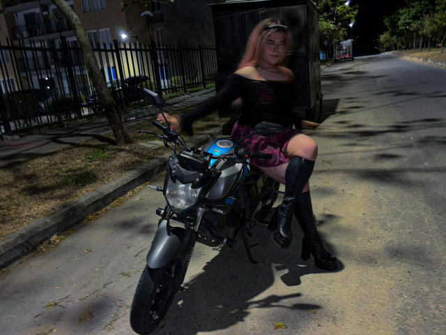 EvelyneRosse with pink hair, black off-shoulder top, and plaid skirt, sitting on a gray motorcycle at night, wearing black boots and a cap, holding the handlebars.