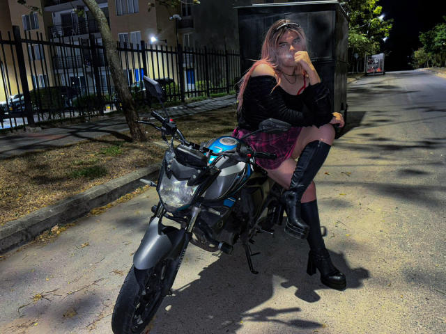 EvelyneRosse with pink hair, black top, pink plaid skirt, black boots, sitting on black motorcycle at night, sucking finger, motorcycle in foreground, urban setting, shadows on pavement.