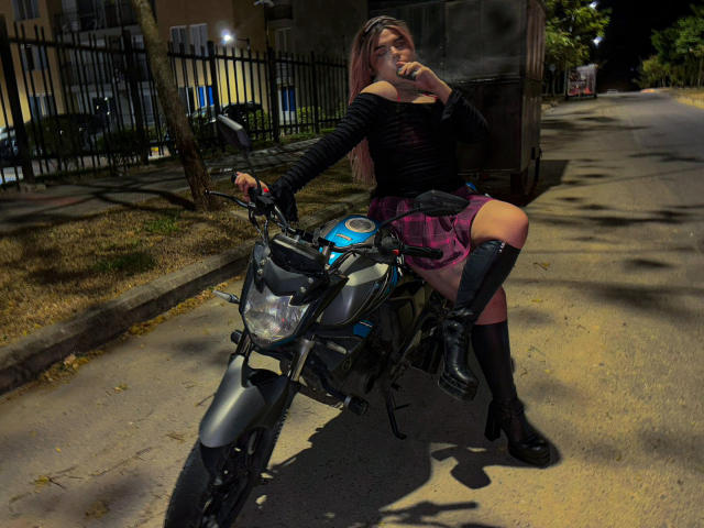 EvelyneRosse with long pink hair, black top, pink plaid skirt, black boots, and knee socks, sitting on a black and blue motorcycle at night, smoking a cigarette.