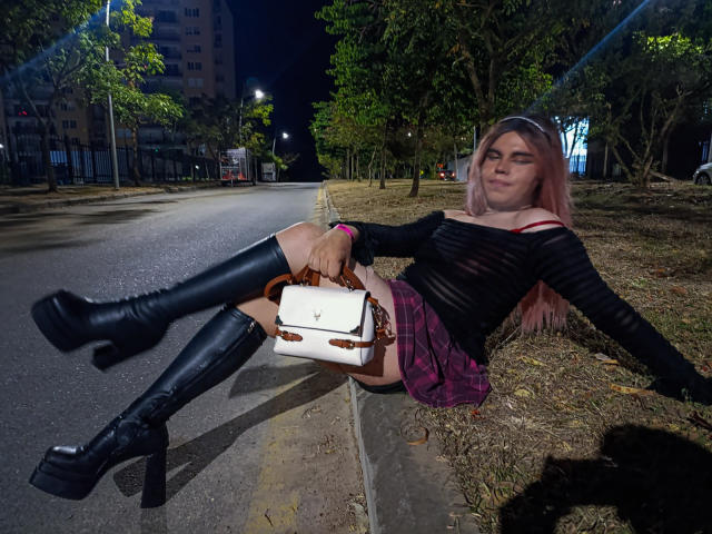 EvelyneRosse with pink hair, black sheer top, plaid skirt, black knee-high boots, and white purse, sitting on sidewalk, legs up, night, urban, street, trees, confident, sexy.