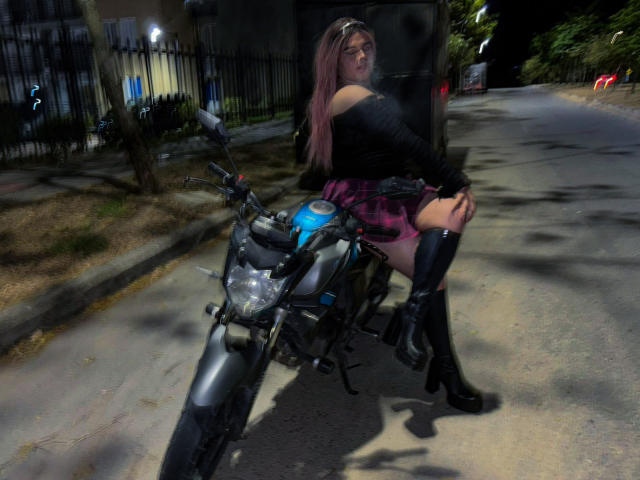 EvelyneRosse with pink hair, black off-shoulder top, pink plaid skirt, black knee-high boots, sitting on blue motorcycle, night, sidewalk, shadow, hands on bike, sexy.