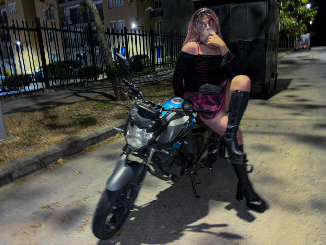 EvelyneRosse with pink hair, black off-shoulder top, pink plaid skirt, black boots, and knee-high socks, sitting on a black motorcycle, drinking from a bottle.