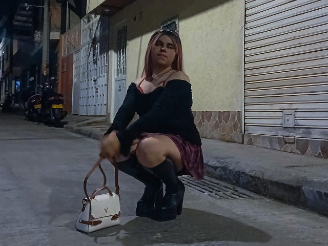 EvelyneRosse, squatting, wearing black off-shoulder top, pink skirt, black socks, platform shoes, holding white purse, street at night, visible ass, medium tits, brown hair, serious expression.