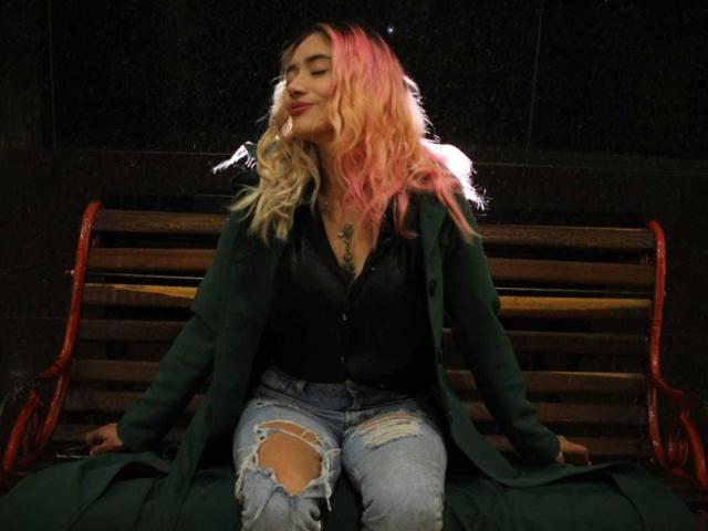 FreankyQueen with pink and blonde wavy hair, green coat, black shirt, and ripped jeans, sitting on a red and wooden bench, eyes closed, smiling.