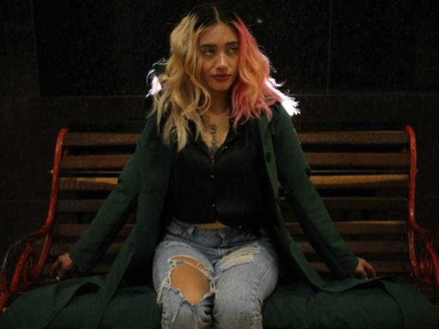 FreankyQueen with blonde and pink wavy hair, wearing a green jacket, black shirt, and ripped jeans, sitting on a red wooden bench at night.
