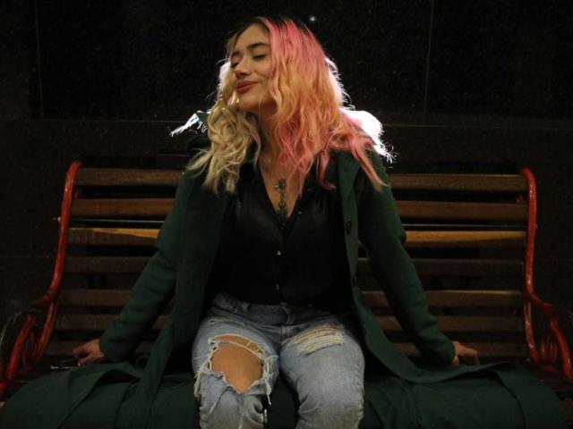 FreankyQueen with pink and blonde hair, green coat, black shirt, and ripped jeans, sitting on a wooden bench with red metal arms, smiling with eyes closed.