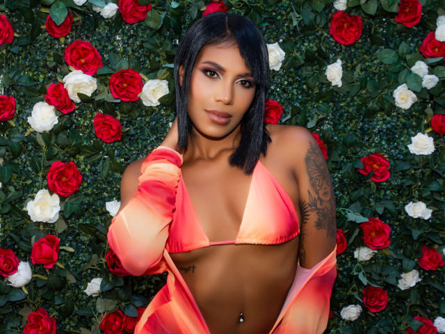 LaylaJones with medium brown skin, black bob, pink-orange bikini top, and matching long-sleeve top, showing tits and a tattoo on her left arm, standing in front of roses.