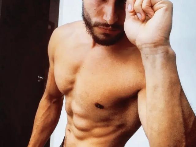 AckeranBigCock, shirtless with a muscular, defined, tan torso, small dark nipples, and a beard, holding his right hand near his face, showing a serious expression.